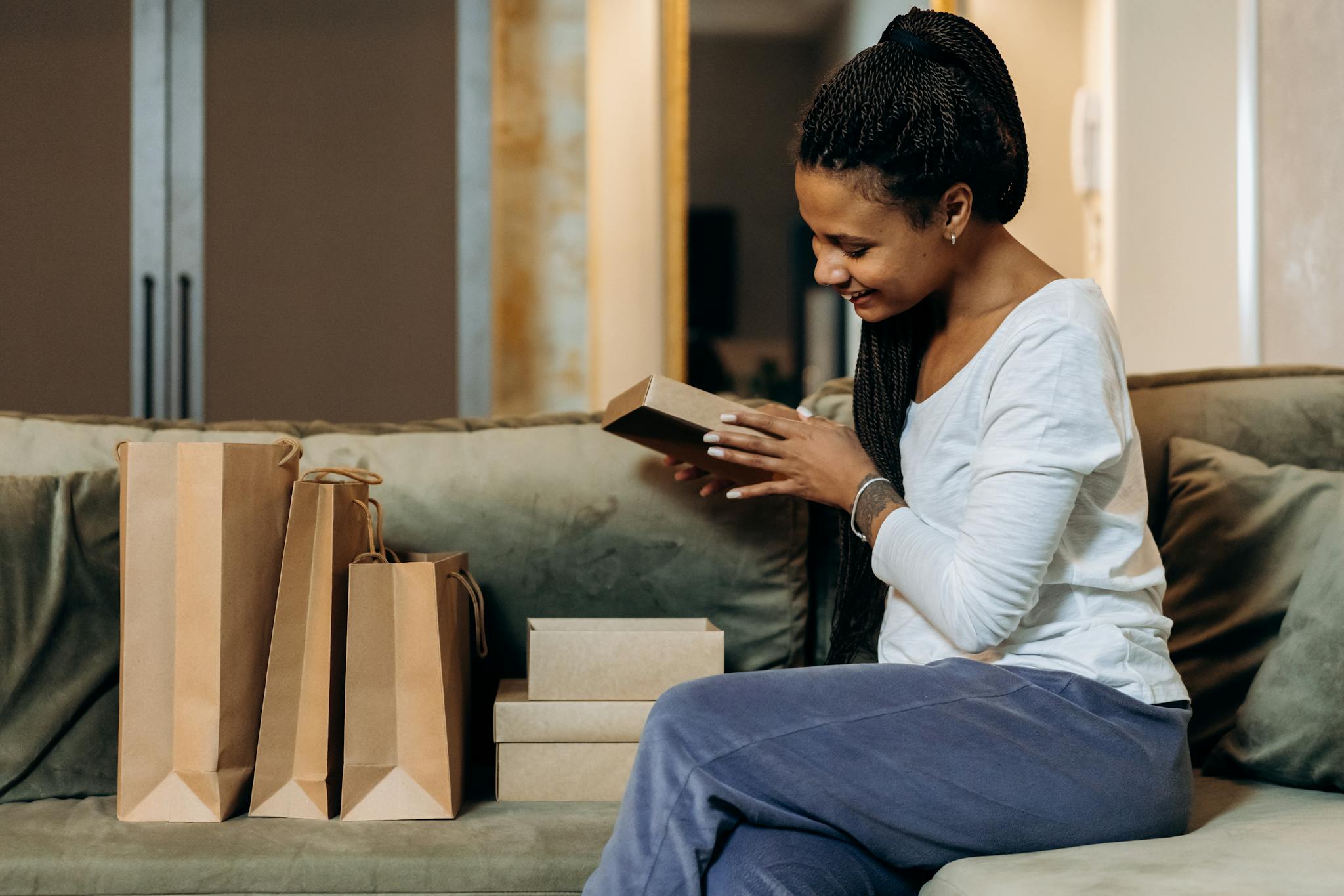 Smiling woman sitting on a couch, unwrapping gifts with paper bags around her.